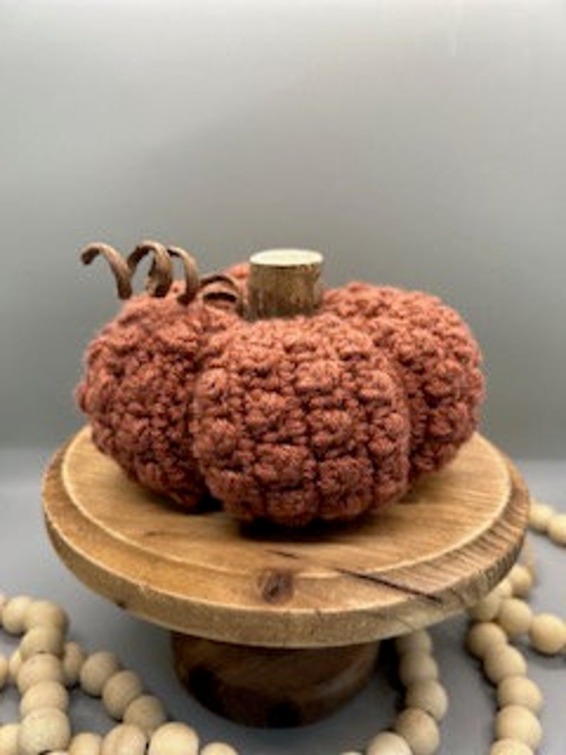 May include: A crocheted pumpkin in a terracotta colour with a brown wooden stem. The pumpkin is sitting on a wooden pedestal.