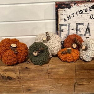 May include: A collection of seven crocheted pumpkins in various colours, including orange, green, and beige, with wooden stems. The pumpkins are arranged on a wooden surface in front of a sign that reads "Antique & Open Daily Flea Market".