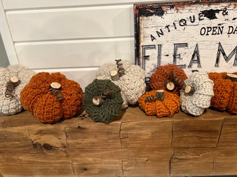 May include: A collection of seven crocheted pumpkins in various colours, including orange, green, and beige, with wooden stems. The pumpkins are arranged on a wooden surface in front of a sign that reads "Antique & Open Daily Flea Market".