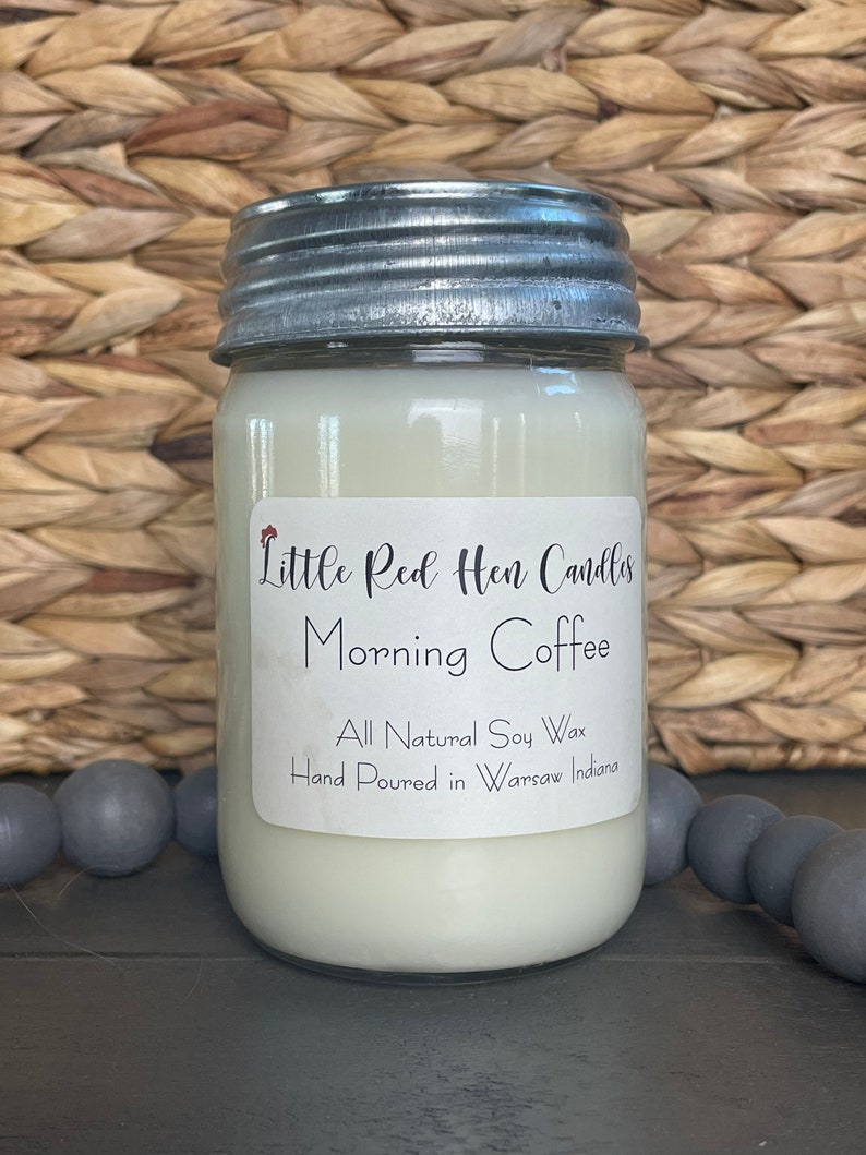 May include: A glass jar candle with a silver lid. The label reads "Little Red Hen Candles Morning Coffee All Natural Soy Wax Hand Poured in Warsaw Indiana."