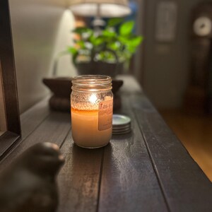 Puede incluir: Una vela encendida en un frasco de vidrio transparente con una etiqueta que dice "Fall" sobre una mesa de madera. La vela est&aacute; rodeada de algunas tapas.
