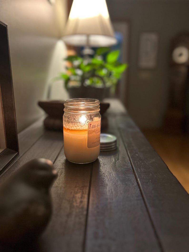 Puede incluir: Una vela encendida en un frasco de vidrio transparente con una etiqueta que dice "Fall" sobre una mesa de madera. La vela est&aacute; rodeada de algunas tapas.