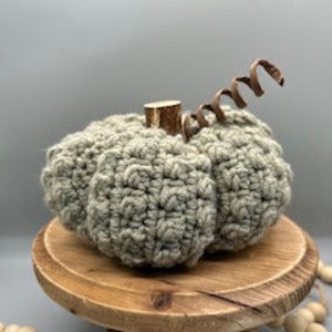 May include: A crocheted green pumpkin with a brown wooden stem. The pumpkin is sitting on a wooden pedestal with a string of wooden beads in the background.