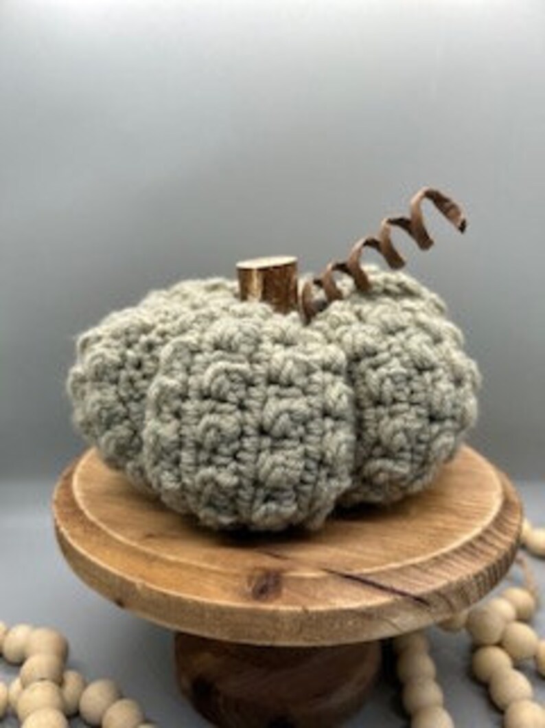 May include: A crocheted green pumpkin with a brown wooden stem. The pumpkin is sitting on a wooden pedestal with a string of wooden beads in the background.
