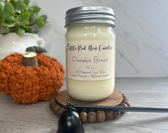 Op de afbeelding: Een kaars van 340 gram in een glazen pot met een zilveren deksel. De kaars is gelabeld met "Little Red Hen Candles" en "Pumpkin Bread". De kaars staat op een houten onderzetter met een bruin en zwart ontwerp.