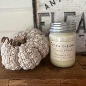 May include: A glass jar candle with a silver lid. The candle is labelled "Little Pie Hot Cocoa" and "Pumpkin Bread". The candle is sitting on a wooden surface next to a crocheted pumpkin.