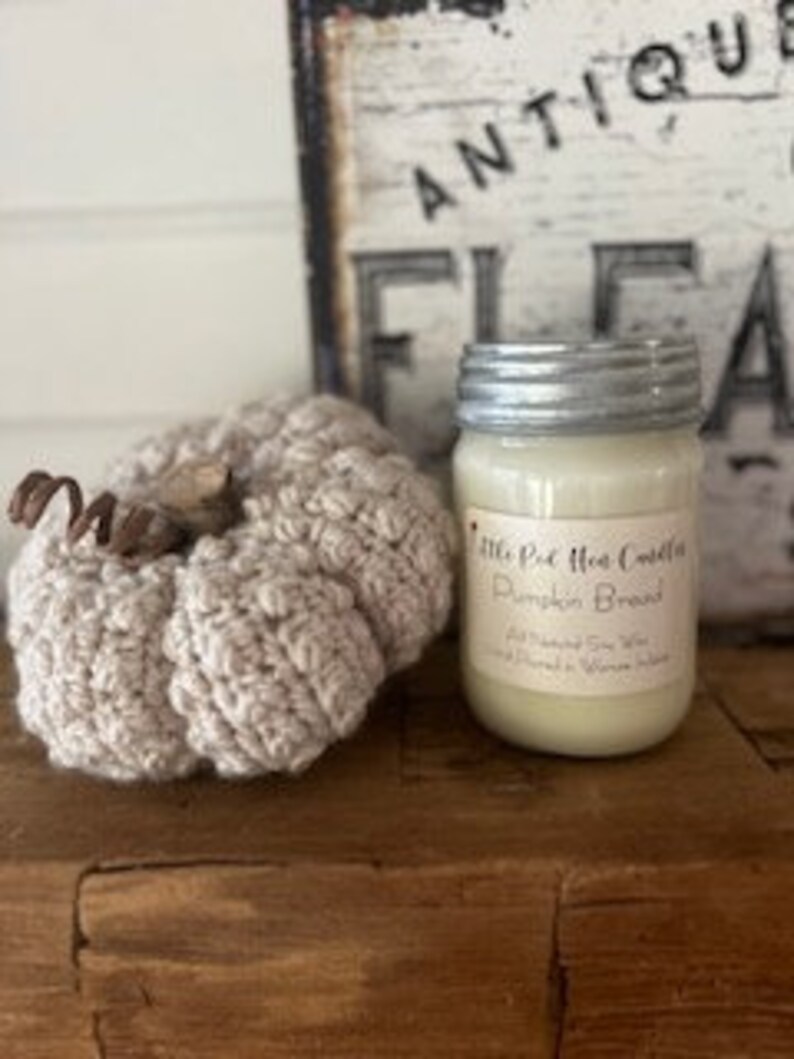 May include: A glass jar candle with a silver lid. The candle is labelled "Little Pie Hot Cocoa" and "Pumpkin Bread". The candle is sitting on a wooden surface next to a crocheted pumpkin.