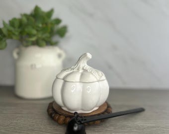 Op de afbeelding: Witte keramische pompoenvormige container met een deksel. De container staat op een houten onderzetter met een zwart metalen handvat.