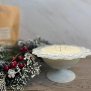 Op de afbeelding: Een witte kaars in een vintage-stijl voetkom met een gegolfde rand. De kaars is omringd door een besneeuwde groenblijvende krans met rode bessen.