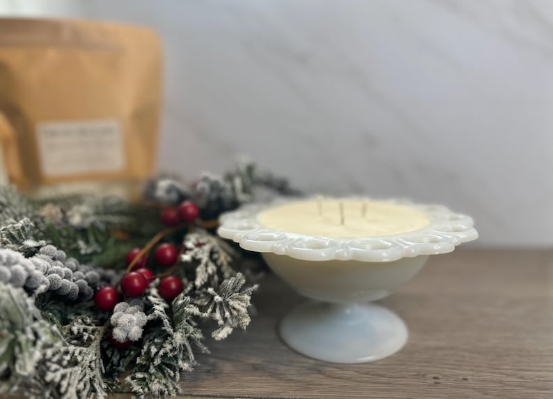 Op de afbeelding: Een witte kaars in een vintage-stijl voetkom met een gegolfde rand. De kaars is omringd door een besneeuwde groenblijvende krans met rode bessen.