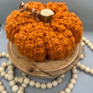 May include: A crocheted orange pumpkin with a brown stem and a hessian bow. The pumpkin is sitting on a wooden stand with white beads.
