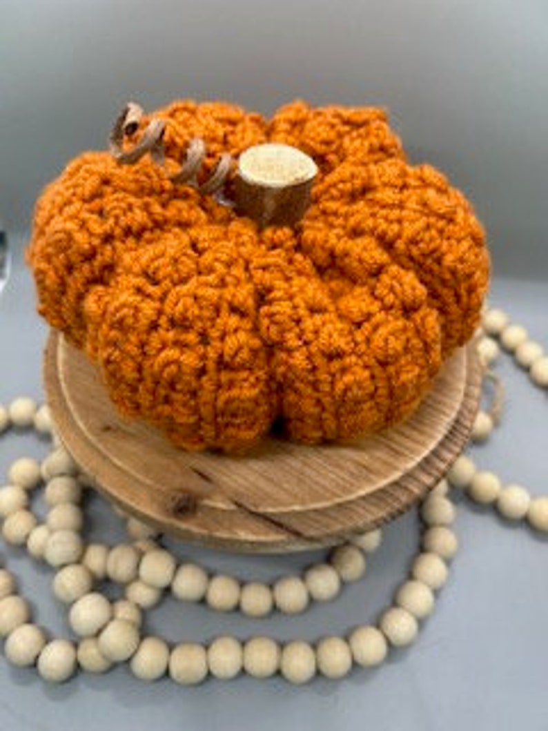 May include: A crocheted orange pumpkin with a brown stem and a hessian bow. The pumpkin is sitting on a wooden stand with white beads.