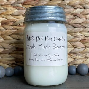 Puede incluir: Una vela blanca en un frasco de vidrio con tapa plateada. La etiqueta dice "Little Red Hen Candles Apple Maple Bourbon All Natural Soy Wax Hand Poured in Warsaw Indiana."