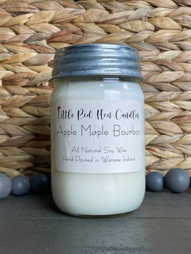 Puede incluir: Una vela blanca en un frasco de vidrio con tapa plateada. La etiqueta dice "Little Red Hen Candles Apple Maple Bourbon All Natural Soy Wax Hand Poured in Warsaw Indiana."