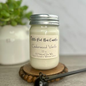 May include: A jar candle with a silver lid, labeled "Little Red Hen Candles Cedarwood Vanilla." The label indicates 12 oz of all-natural soy wax, hand-poured in Warsaw, Indiana. The candle sits on a wooden coaster with a beaded edge.