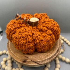May include: A crocheted orange pumpkin with a brown stem. The pumpkin is sitting on a wooden base with a string of wooden beads in the background.