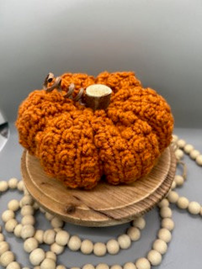 May include: A crocheted orange pumpkin with a brown stem. The pumpkin is sitting on a wooden base with a string of wooden beads in the background.