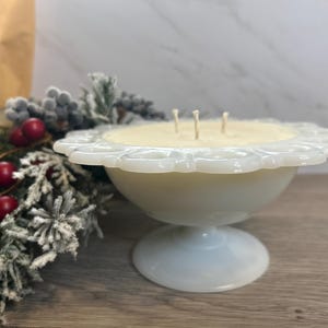 Op de afbeelding: Een witte kaars in een vintage melkglas voetstuk met een gegolfde rand. De kaars heeft drie lonten en is omringd door een sneeuwwitte en rode kerstdecoratie.