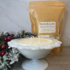 Op de afbeelding: Een witte kaars in een witte, gegolfde kom staat op een houten oppervlak. De kaars is gelabeld "Little Red Hen Candles Banana Nut Bread" en is gemaakt van volledig natuurlijke sojawas. De kaars is met de hand gegoten in Warren, Indiana.