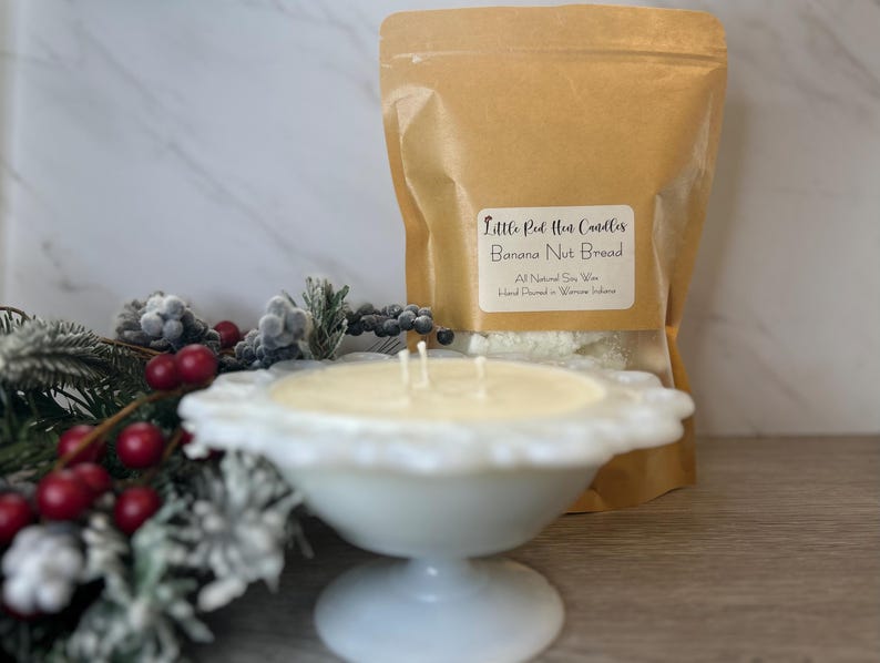 Op de afbeelding: Een witte kaars in een witte, gegolfde kom staat op een houten oppervlak. De kaars is gelabeld "Little Red Hen Candles Banana Nut Bread" en is gemaakt van volledig natuurlijke sojawas. De kaars is met de hand gegoten in Warren, Indiana.