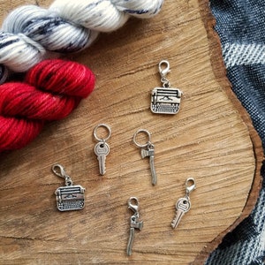 May include: Six silver stitch markers with different charms: a typewriter, a key, an axe, and two more axes. The charms are on a wooden surface with yarn in the background.