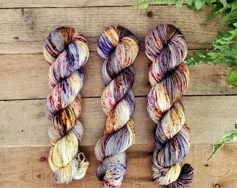 May include: Three skeins of yarn in a variety of colors, including purple, yellow, and brown. The yarn is arranged in a vertical line on a wooden surface.