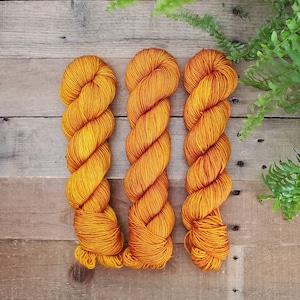 May include: Three skeins of yarn in a bright orange color. The yarn is wound into a ball shape and is ready to be used for knitting or crocheting.