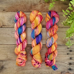 May include: Three skeins of colourful yarn in shades of pink, orange, yellow, blue, and purple. The yarn is arranged in a row on a wooden surface.