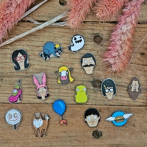 May include: A selection of colourful cartoon character pins, including a ghost, a raccoon with a crown, and several characters with glasses and moustaches. The pins are arranged on a wooden surface with pink dried flowers.
