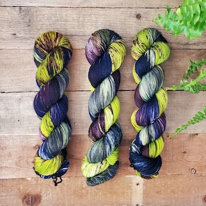 May include: Three skeins of yarn in a dark blue, green, and purple color combination. The yarn is arranged in a vertical line on a wooden surface.
