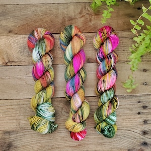 May include: Three skeins of colorful yarn in shades of green, yellow, pink, and black. The yarn is arranged vertically on a wooden surface.