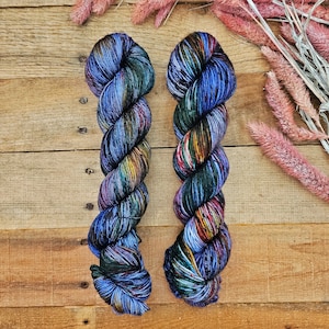 May include: Two skeins of hand-dyed yarn in a vibrant array of colors, including blues, greens, and hints of yellow and red. The yarn is twisted and arranged on a rustic wooden surface, with dried pink flowers in the background. A beautiful fiber art supply.
