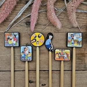 May include: A collection of six decorative cocktail picks featuring cartoon character designs. The picks have various rectangular and circular toppers with colorful illustrations of a family. The picks are on wooden sticks, arranged on a wooden surface.