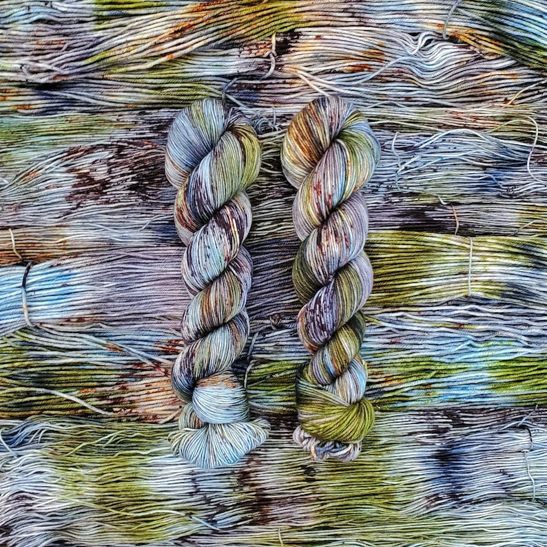 May include: Two skeins of yarn in a variegated colorway with shades of green, blue, brown, and purple. The yarn is arranged in a twisted pattern.