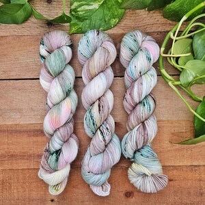 May include: Three twisted skeins of yarn featuring a variegated color palette of soft pastel pink, mint green, light blue, and cream. The yarn displays subtle speckles of darker tones throughout the fibers, creating a multi-colored appearance.