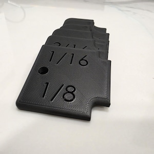 May include: A stack of black 3D printed plastic gauges with a textured surface. Each piece features embossed numerical markings including 1/16 and 1/8, with a circular hole near the top corner and a notched edge on the right side of each unit.