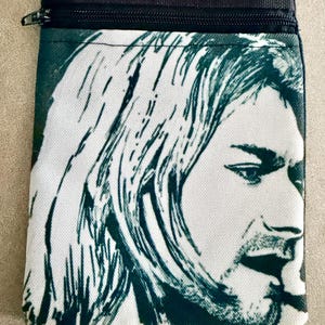 May include: A black and white crossbody bag featuring a high-contrast graphic print of a person with long hair on the front panel. The bag includes two horizontal zippered compartments at the top and a black adjustable strap attached to the upper corners.