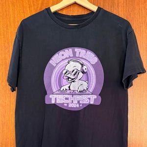 May include: A black short-sleeve t-shirt featuring a purple circular graphic on the chest. The graphic depicts a cartoon duck wearing headphones and sunglasses, DJing at a console. The text reads NEON TREES TECHFEST 2024 in white lettering.