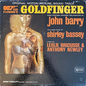 May include: Goldfinger soundtrack album cover. The album features a gold-painted woman, with a couple in the center. The text includes "Original Motion Picture Sound Track", "John Barry", and "Shirley Bassey". The album is a high fidelity record.