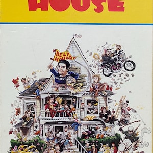 May include: A vintage VHS tape cover for the movie "National Lampoon's Animal House". The cover features a cartoon illustration of a large house with a party scene, including people, animals, and a car. The title "National Lampoon's Animal House" is written in red and yellow letters.