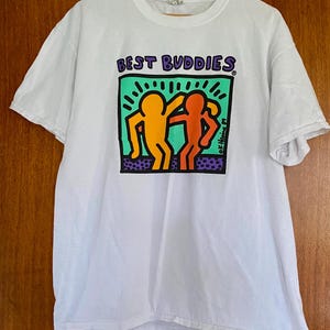 Puede incluir: Camiseta blanca con la frase "BEST BUDDIES" en morado sobre un gr&aacute;fico de dos figuras abraz&aacute;ndose. Las figuras son de color naranja y rojo, sobre un fondo verde y morado. La camiseta est&aacute; colgada de una percha de madera.