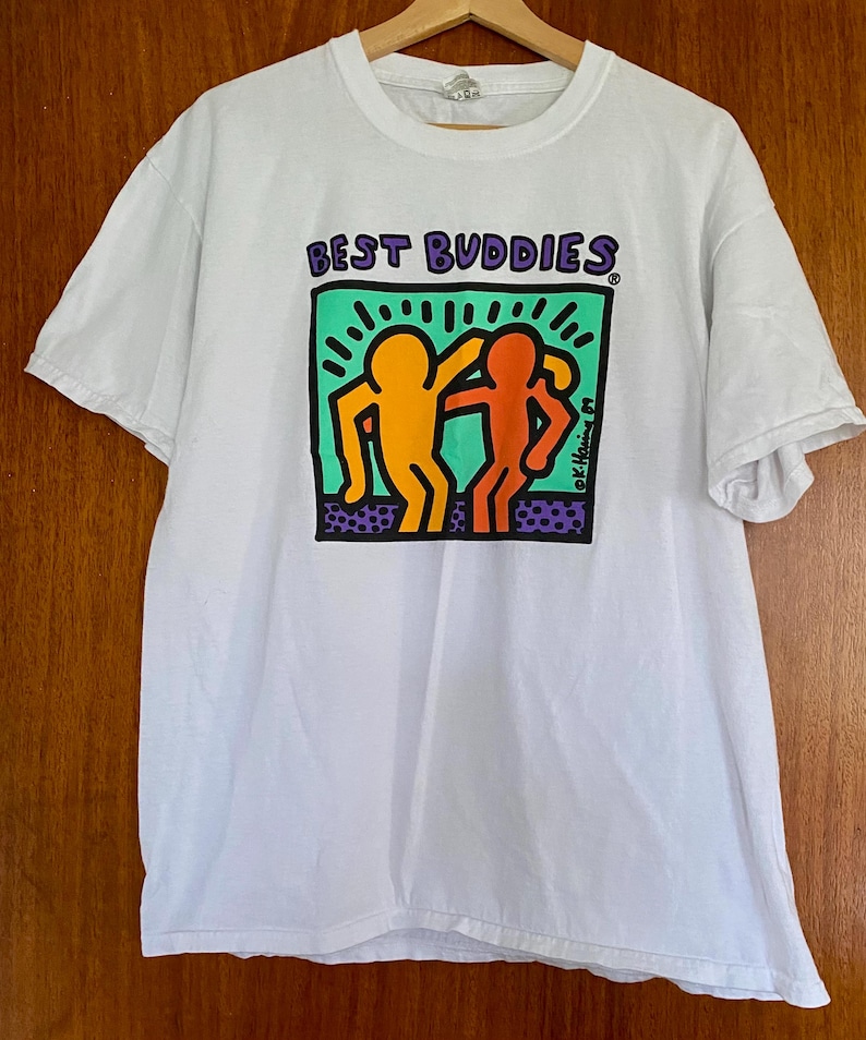 Puede incluir: Camiseta blanca con la frase "BEST BUDDIES" en morado sobre un gr&aacute;fico de dos figuras abraz&aacute;ndose. Las figuras son de color naranja y rojo, sobre un fondo verde y morado. La camiseta est&aacute; colgada de una percha de madera.