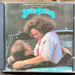 May include: A CD case for the "Sweet Dreams" soundtrack. The cover features a couple embracing in a car, with the title in turquoise script. The text reads "ORIGINAL MOTION PICTURE SOUNDTRACK" and "THE LIFE AND TIMES OF PATSY CLINE."