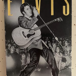 May include: A VHS tape titled "The Great Performances Elvis Center Stage." The cover features a black and white image of Elvis Presley singing and playing a guitar on stage, with a crowd in the background. The title is in gold lettering.