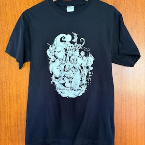 May include: Black t-shirt with a light blue graphic design. The graphic features a collection of cartoonish characters and shapes. The shirt is hanging on a wooden hanger.