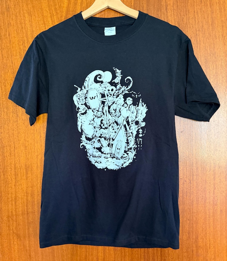 May include: Black t-shirt with a light blue graphic design. The graphic features a collection of cartoonish characters and shapes. The shirt is hanging on a wooden hanger.