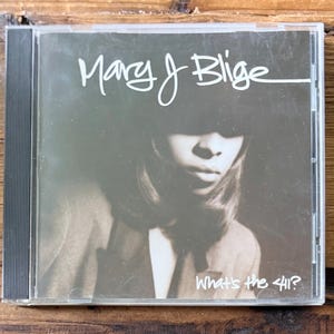 May include: A CD case featuring the album "What's the 411?" by Mary J. Blige. The cover has a sepia-toned image of a person wearing a hat. The artist's name is written in cursive script above the image, and the album title is below.