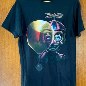 May include: Black t-shirt featuring a colorful graphic of a character with a balloon. The character has a round head with a striped hat, a single blue eye, and a wide smile. A dragonfly is on top of the head.