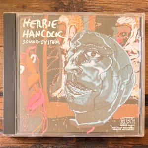 May include: A Herbie Hancock "Sound-System" CD in a clear jewel case. The album art features a stylized portrait in shades of gray and tan. The CD case has a gray spine and the Compact Disc logo is visible.