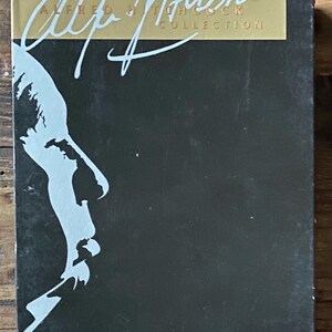 May include: A black box set featuring the title "The Alfred Hitchcock Collection" in gold and white lettering. A stylized, light blue silhouette of a man's profile is on the left side of the box. The box is on a wooden surface.